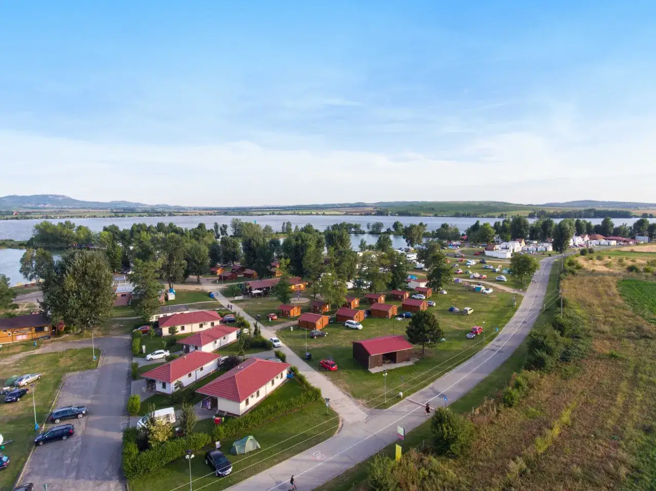 Ferienidylle am See: Kleine H&auml;user und Zelte auf dem Kemp Merkur, umgeben von gr&uuml;nen B&auml;umen und blauer Weite.