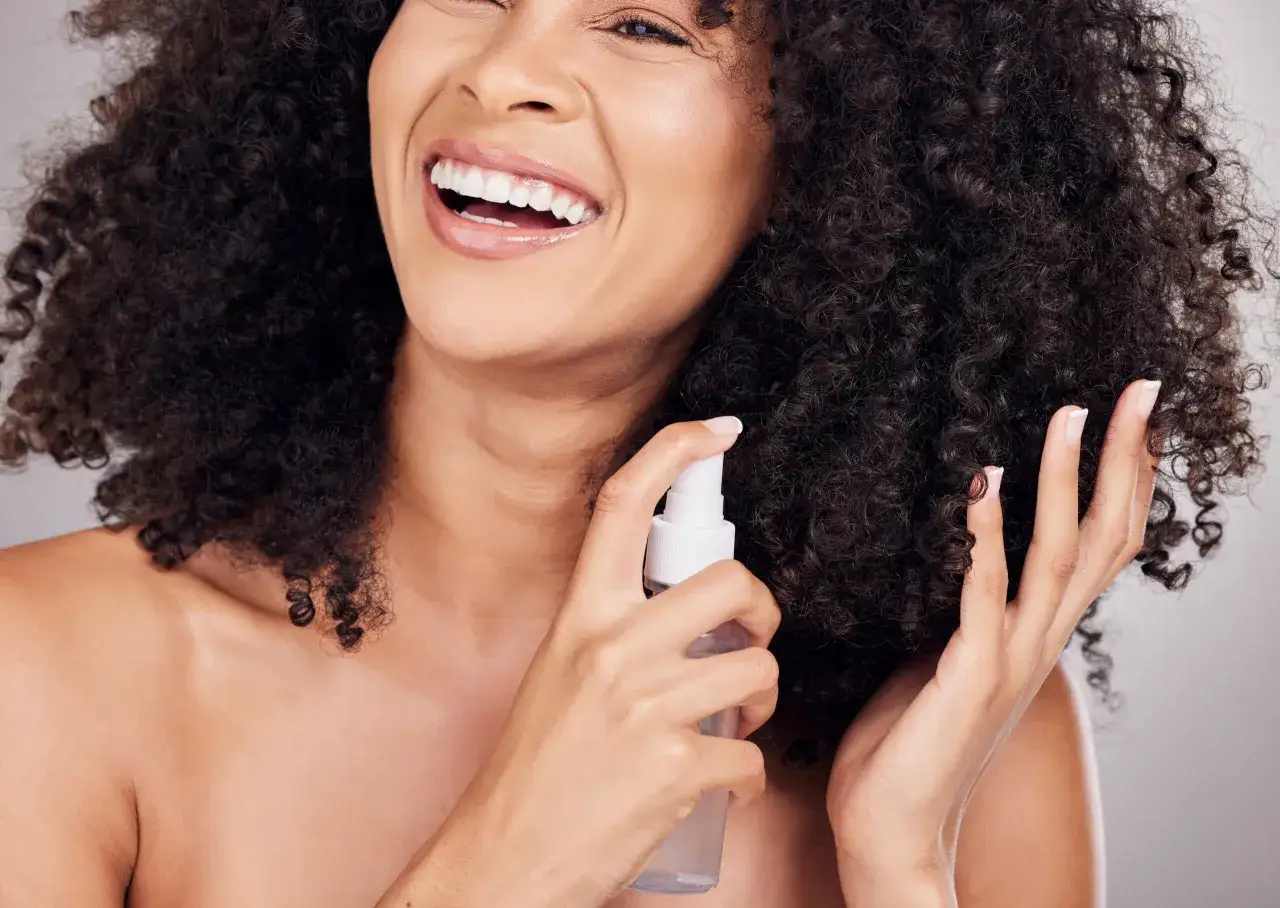 Mujer sonríe mientras aplica spray en su cabello rizado. Aprende como rizar el pelo con espuma para un look espectacular.