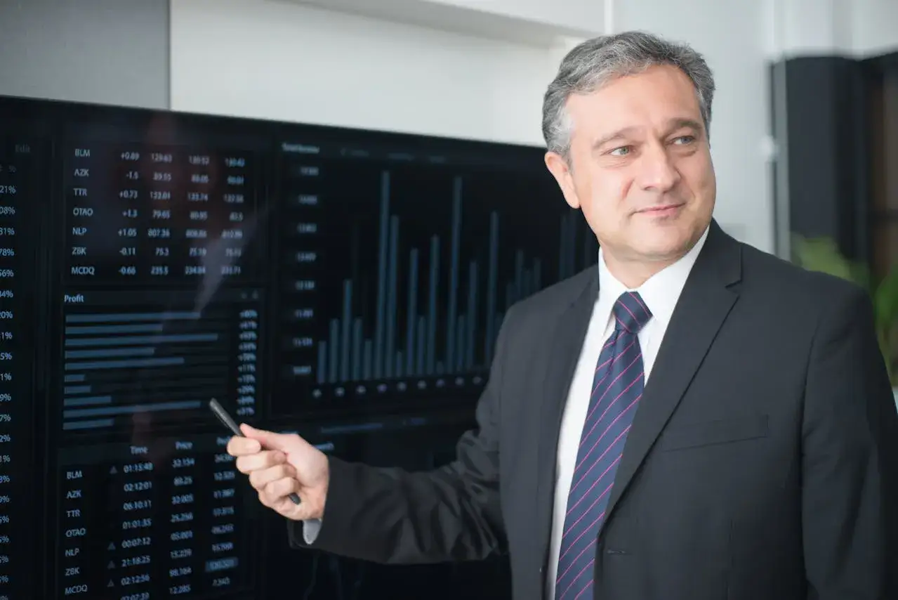 Mężczyzna w garniturze wskazuje na ekran z danymi finansowymi i wykresami.