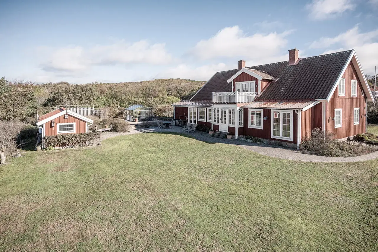 Rött hus med vita knutar och ett litet rött uthus. Gräsmattan är grön och inbjuder till att måla om huset.