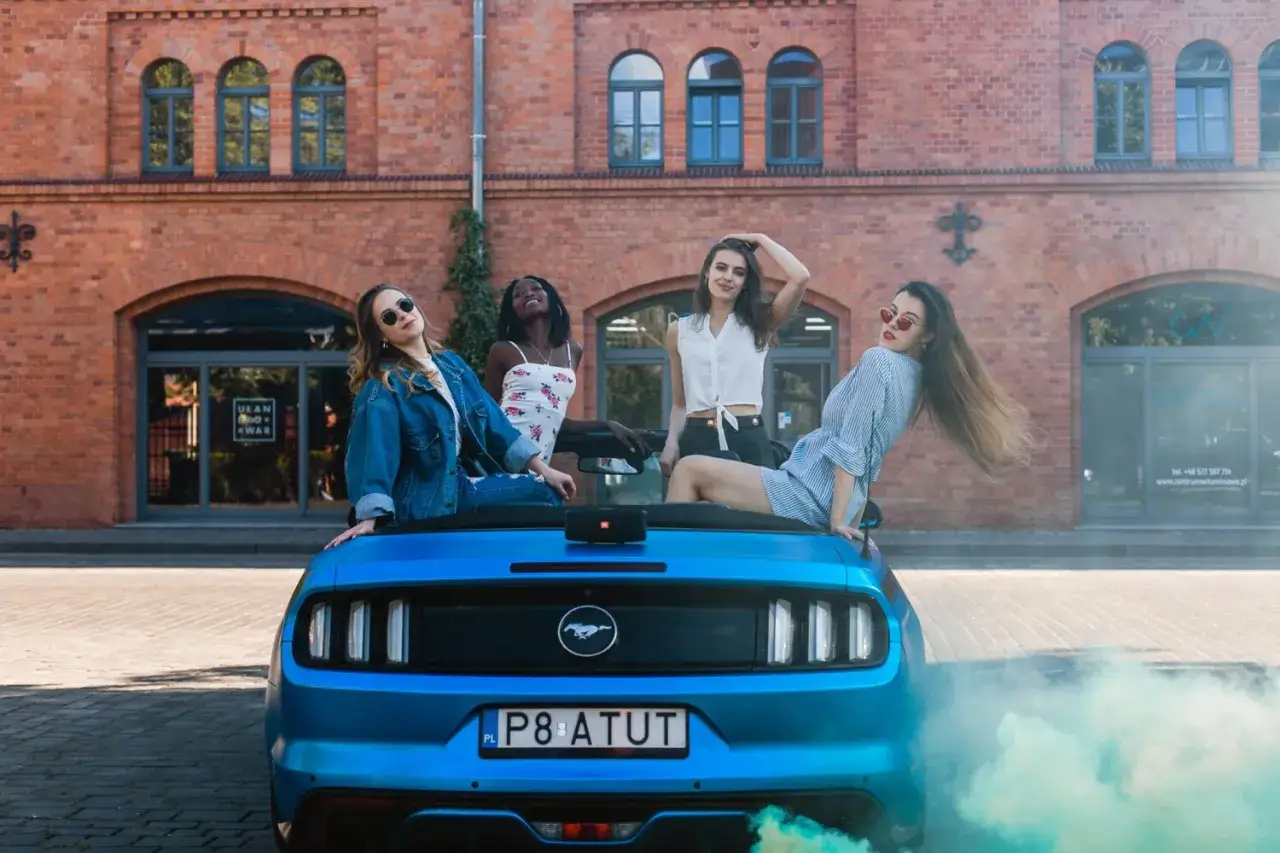 Cztery dziewczyny pozują na tle ceglanego budynku, siedząc na niebieskim mustangu cabrio. Kolorowy dym dodaje atmosfery.