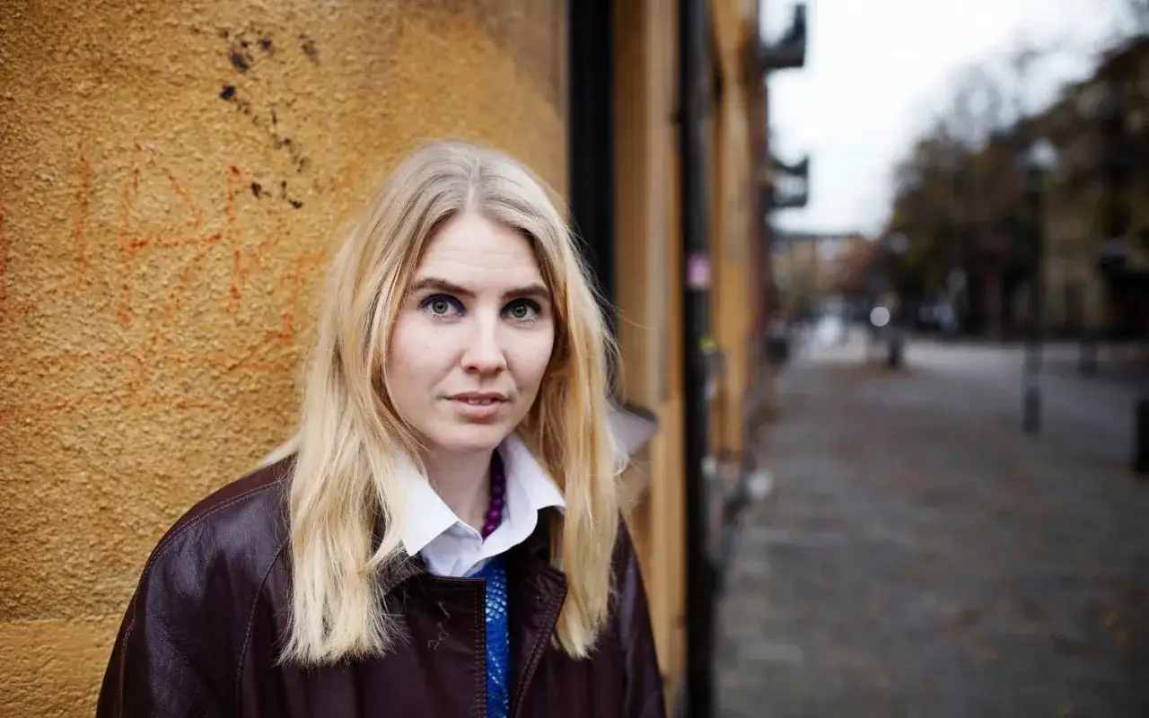 Amanda Romare Lilleman, med sitt blonda hår och blå ögonskugga, står framför en gul vägg.