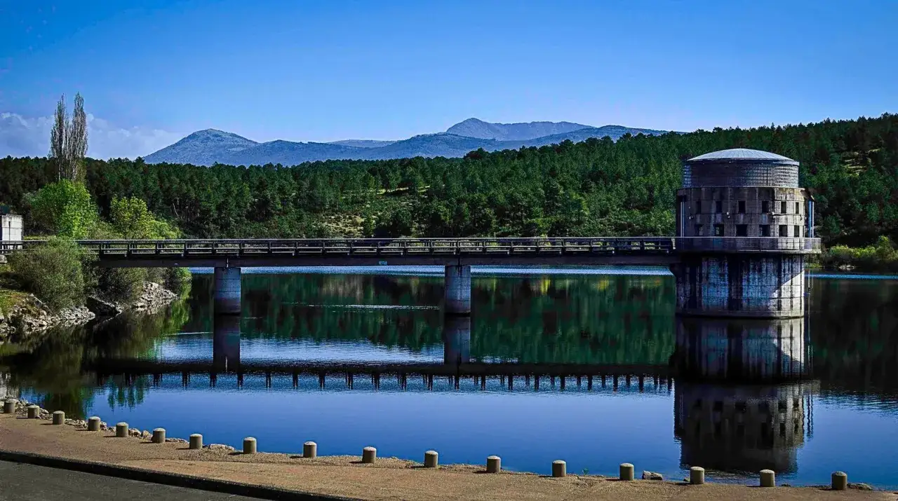 El embalse de Puentes Viejas, con su torre circular y un puente reflejándose en el agua serena, rodeado de bosques y montañas.