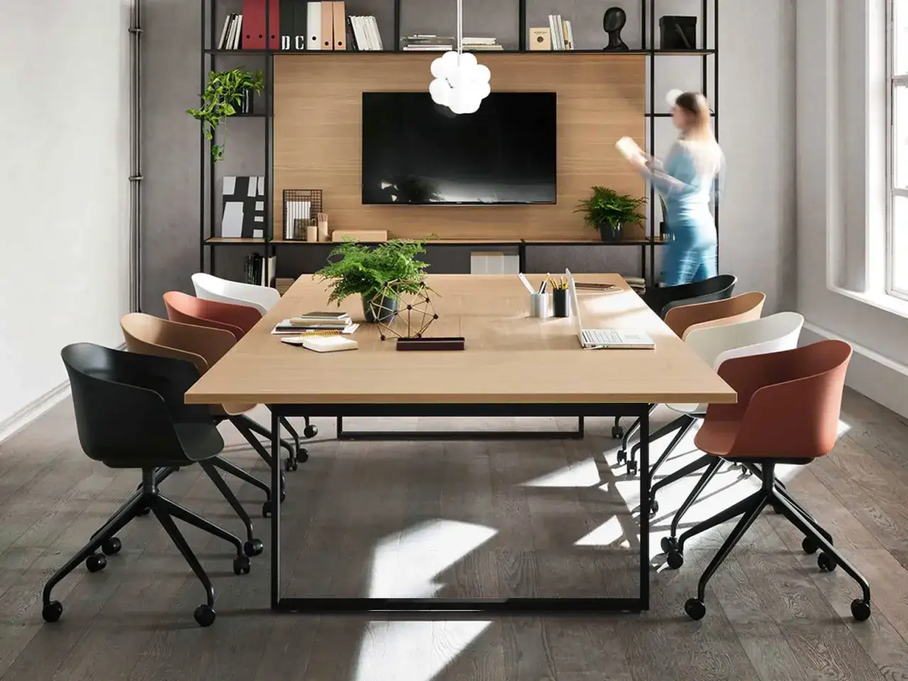 Am&eacute;nagement salle de r&eacute;union moderne avec table en bois, chaises color&eacute;es et &eacute;tag&egrave;res murales. Une femme se tient pr&egrave;s d'un &eacute;cran.