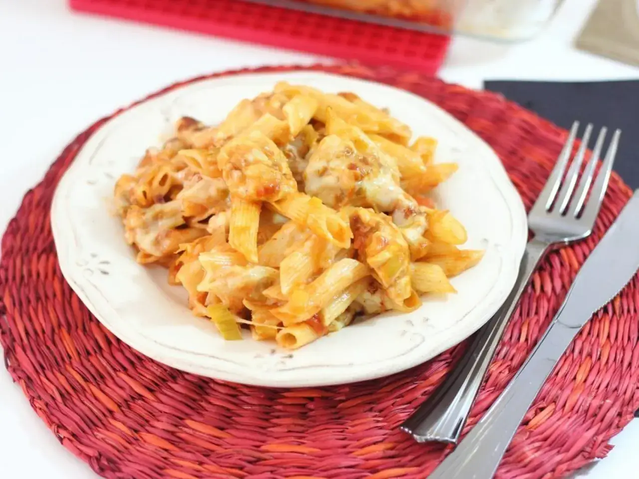 Plato de macarrones con pollo gratinados, cubiertos de queso derretido y salsa roja, servido en un plato blanco sobre una esterilla roja.