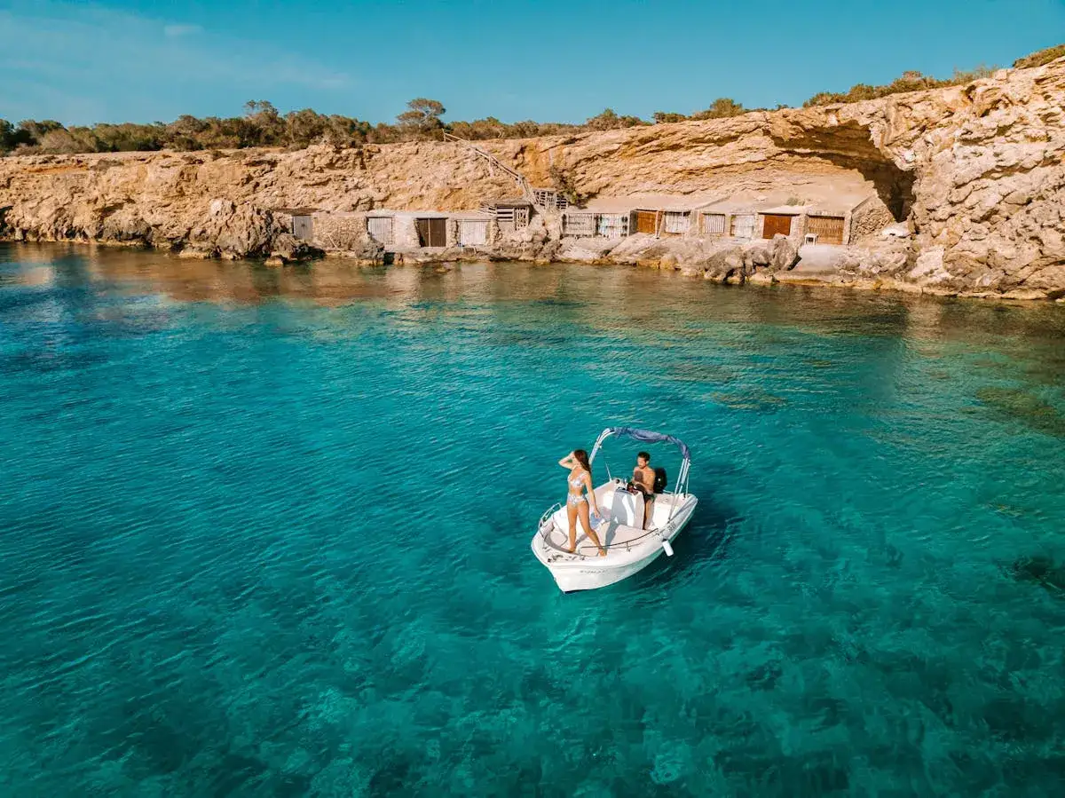 Traumpaar auf einem Boot vor einer felsigen Küste mit kleinen Hütten. Ein idyllischer ibiza strand mit kristallklarem Wasser.
