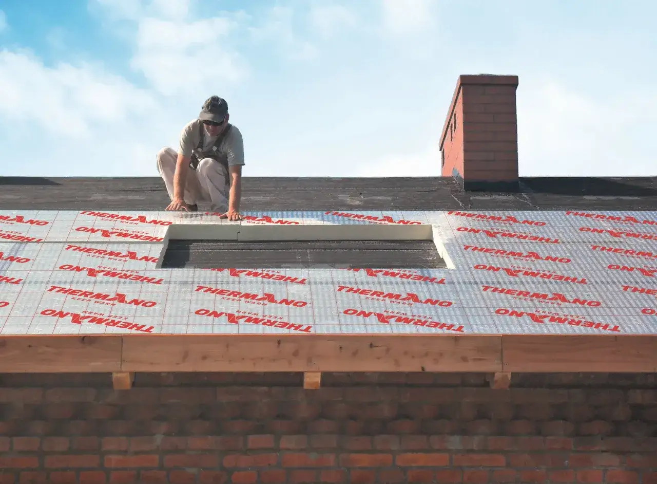 Pracownik układa izolację na dachu, pokazując jak najlepiej ocieplić strop. Widać materiał z logo THERMANO.