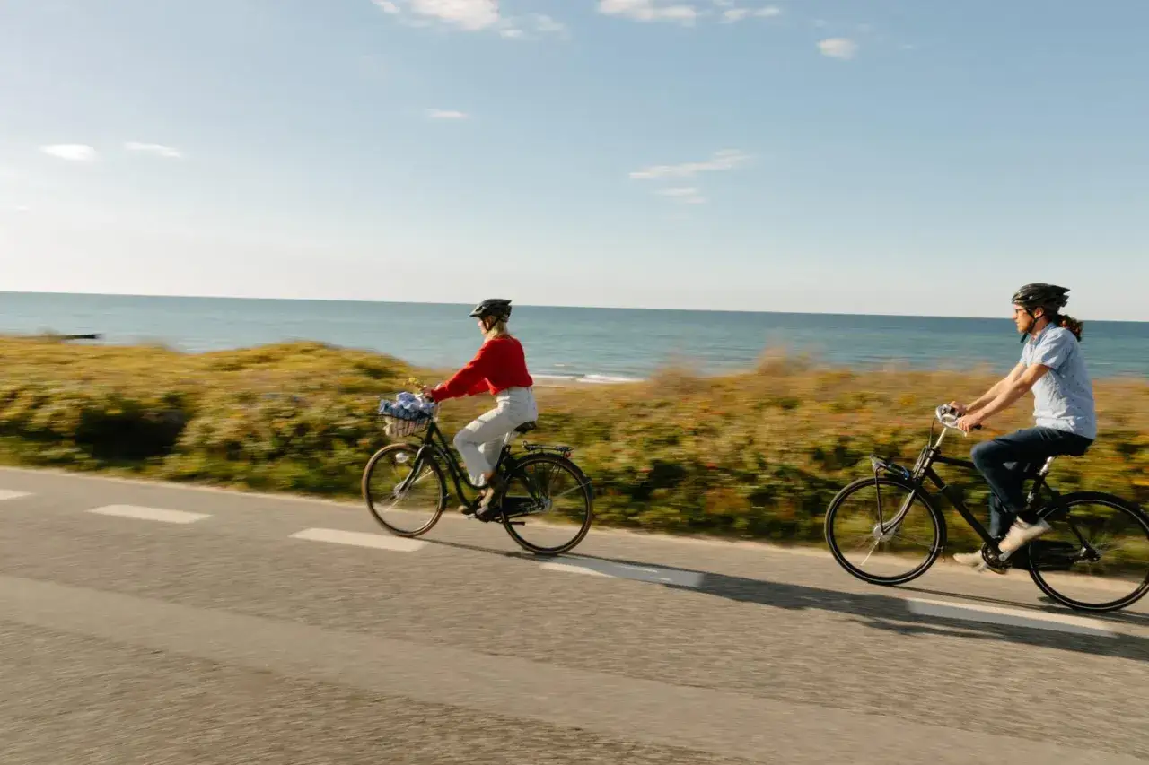 Tv&aring; personer cyklar l&auml;ngs en kustv&auml;g med havet i bakgrunden. En kvinna med en r&ouml;d tr&ouml;ja och en man med en bl&aring; skjorta njuter av cykelturen, kanske p&aring; **cykelleder Danmark**.