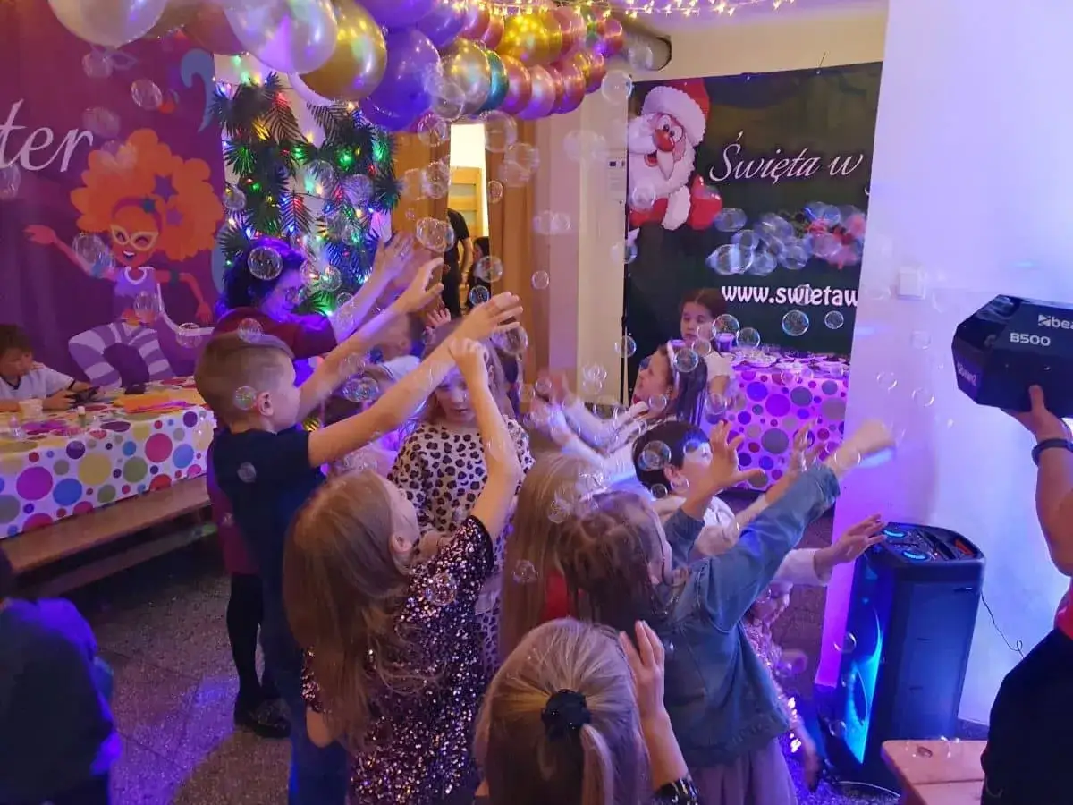 Radosny sylwester dla rodzin z dziećmi: dzieci łapią bańki mydlane, w tle balony i Mikołaj.
