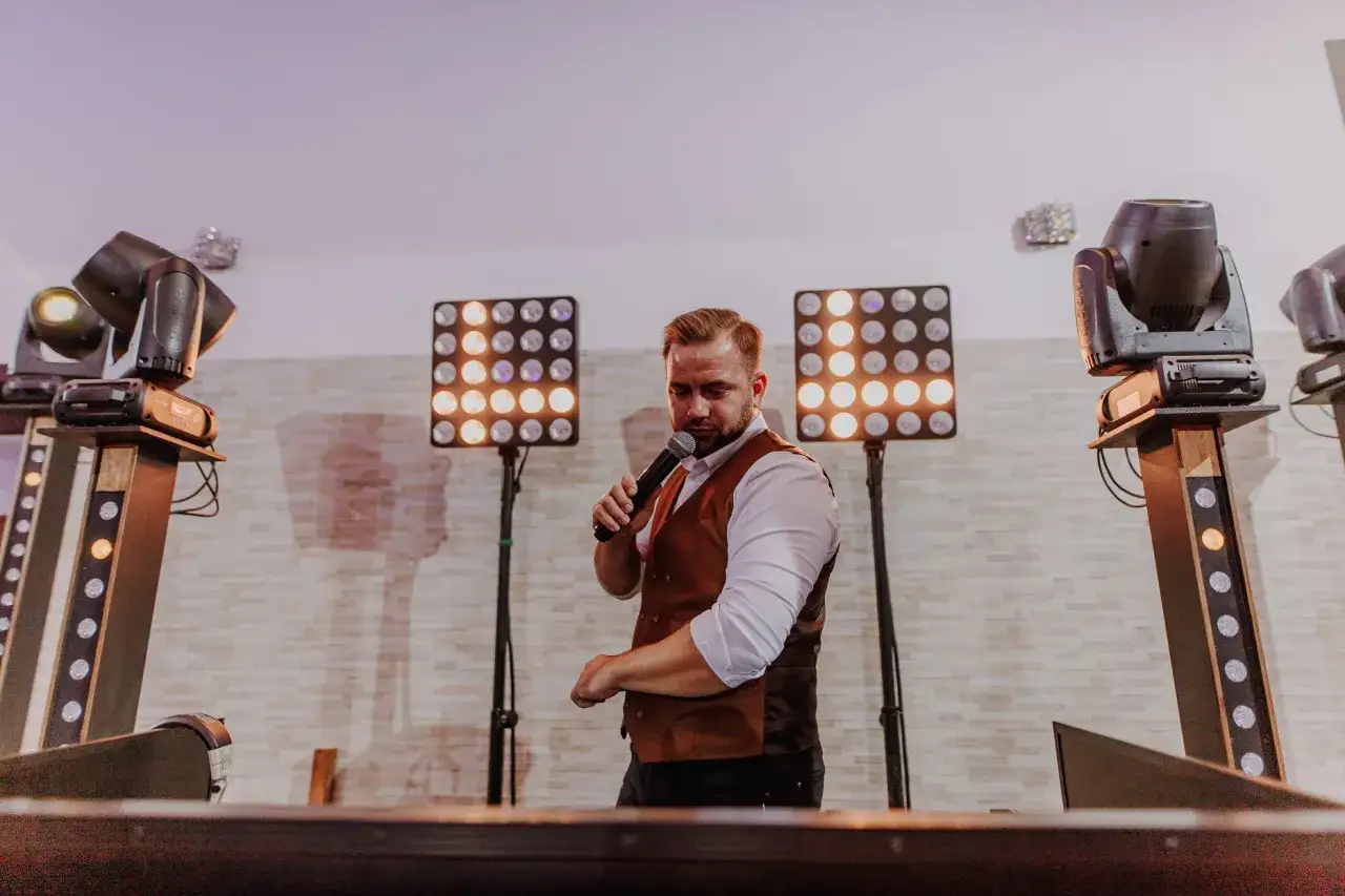 DJ na 18. urodziny, w białej koszuli i brązowej kamizelce, trzyma mikrofon. Za nim świecą panele świetlne i reflektory.