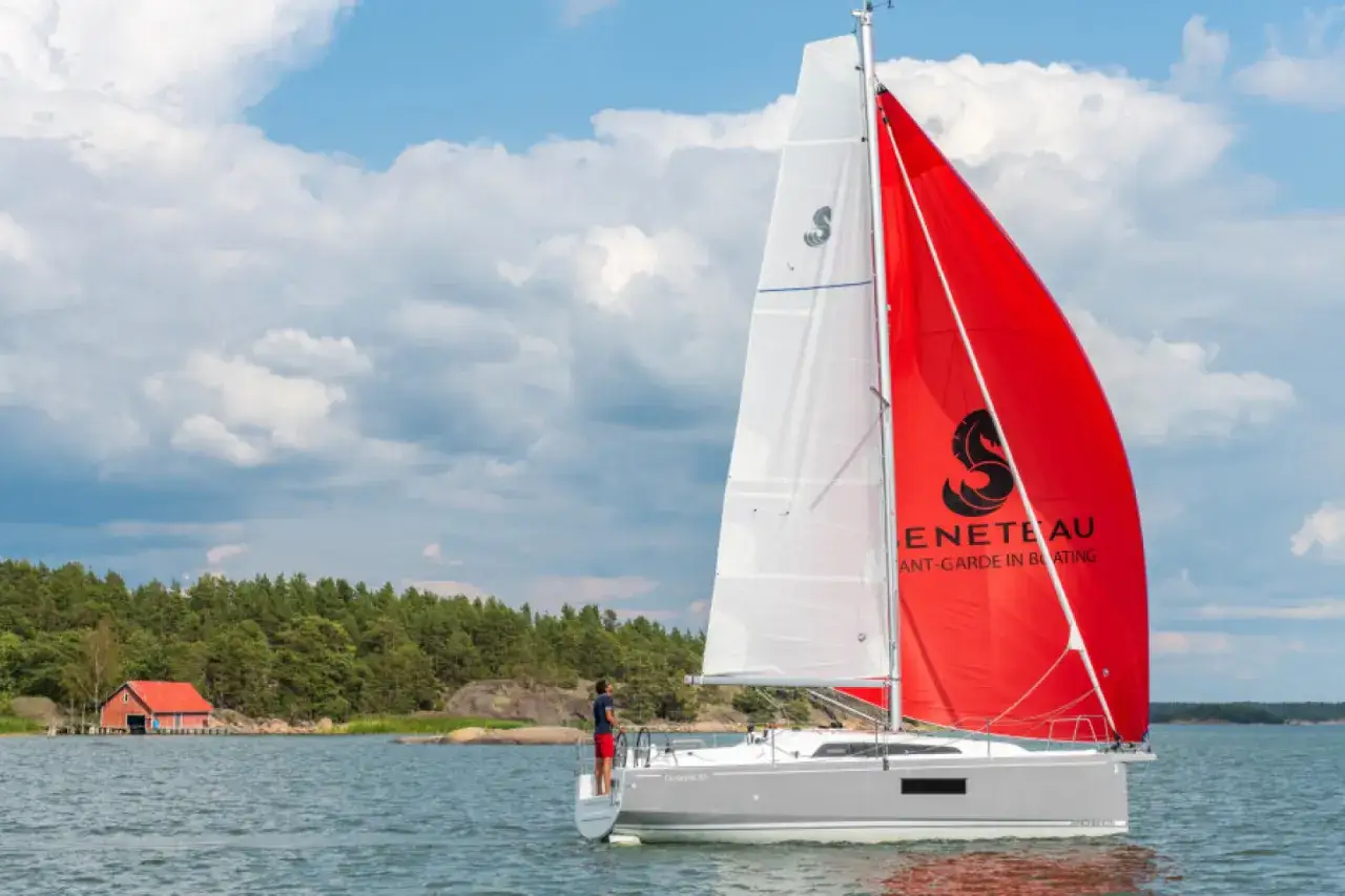 Voilier gris avec une voile rouge et blanche, naviguant près d'une île boisée. Le premier 27.7 avis est prometteur.