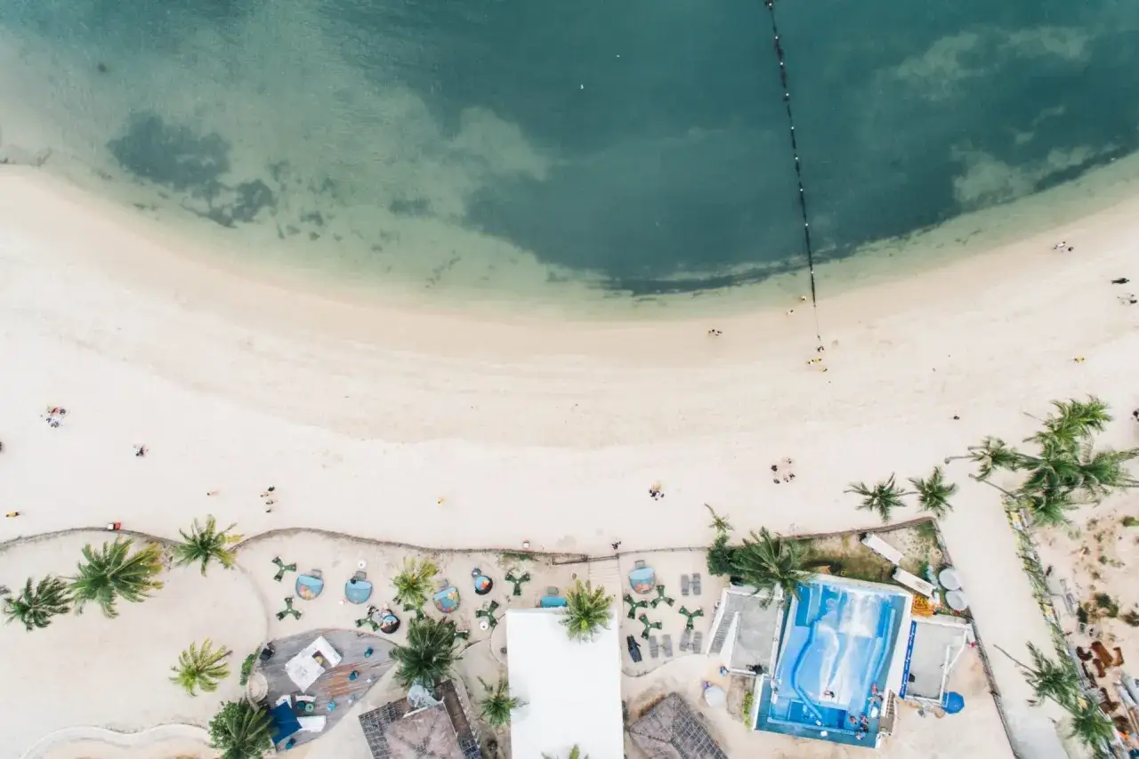 Luftaufnahme eines **Singapur Strandes** mit Palmen, Liegestühlen und einem Wellenbad. Menschen entspannen sich am Sand und im Wasser.
