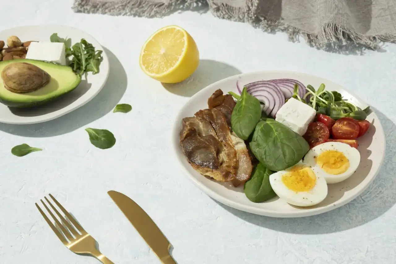 Na talerzu składniki na dietę keto: jajka, boczek, ser feta, szpinak, pomidorki, cebula, awokado i orzechy.