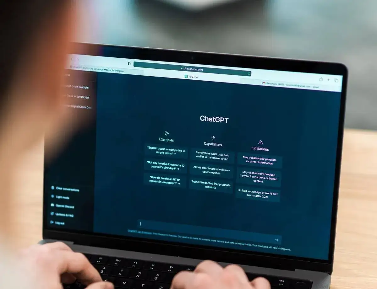 Osoba pisze na klawiaturze laptopa, na ekranie którego widać interfejs chat gpot z przykładami pytań i informacjami o możliwościach.