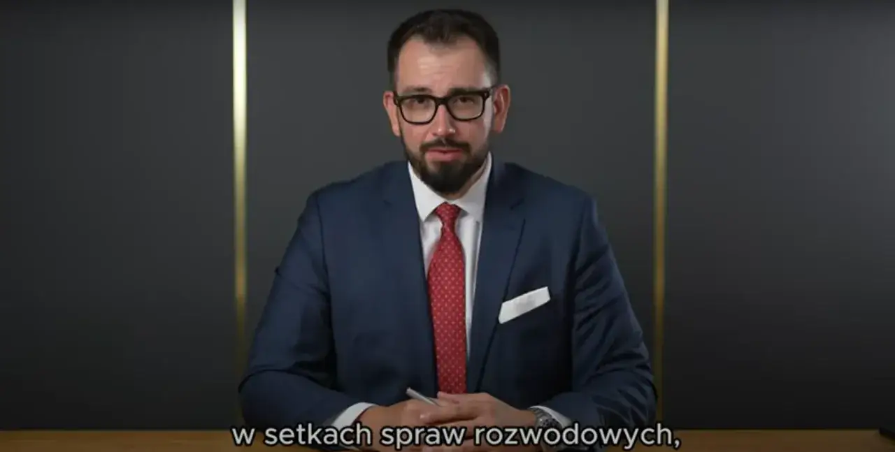 Mężczyzna w garniturze i okularach siedzi przy biurku, trzymając długopis.