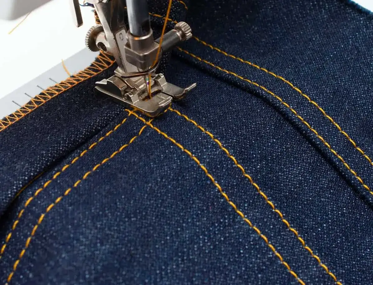 Close-up of a sewing machine needle stitching yellow thread onto dark denim. The presser foot is down, and the stitch is perfectly formed, indicating proper adjusting tension on sewing machine.