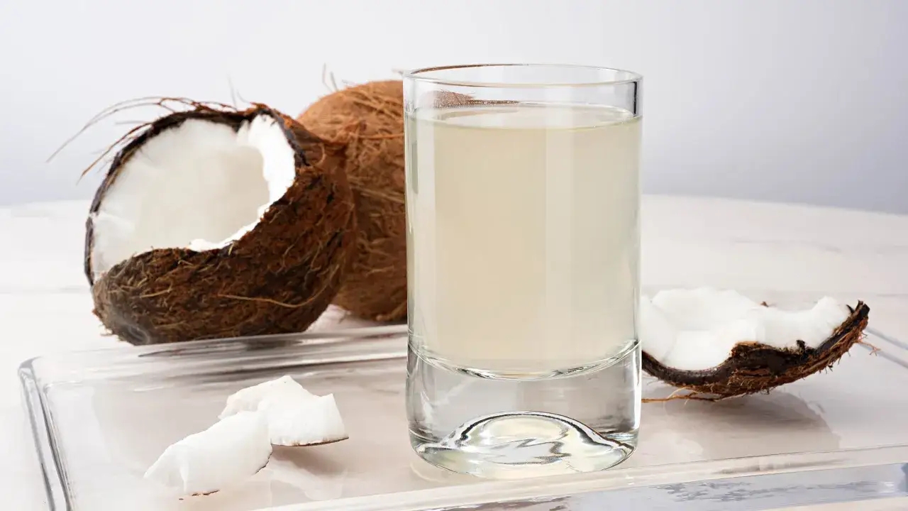 Un vaso de agua de coco fresco junto a cocos partidos. Descubre los **agua de coco beneficios** para tu salud y bienestar.