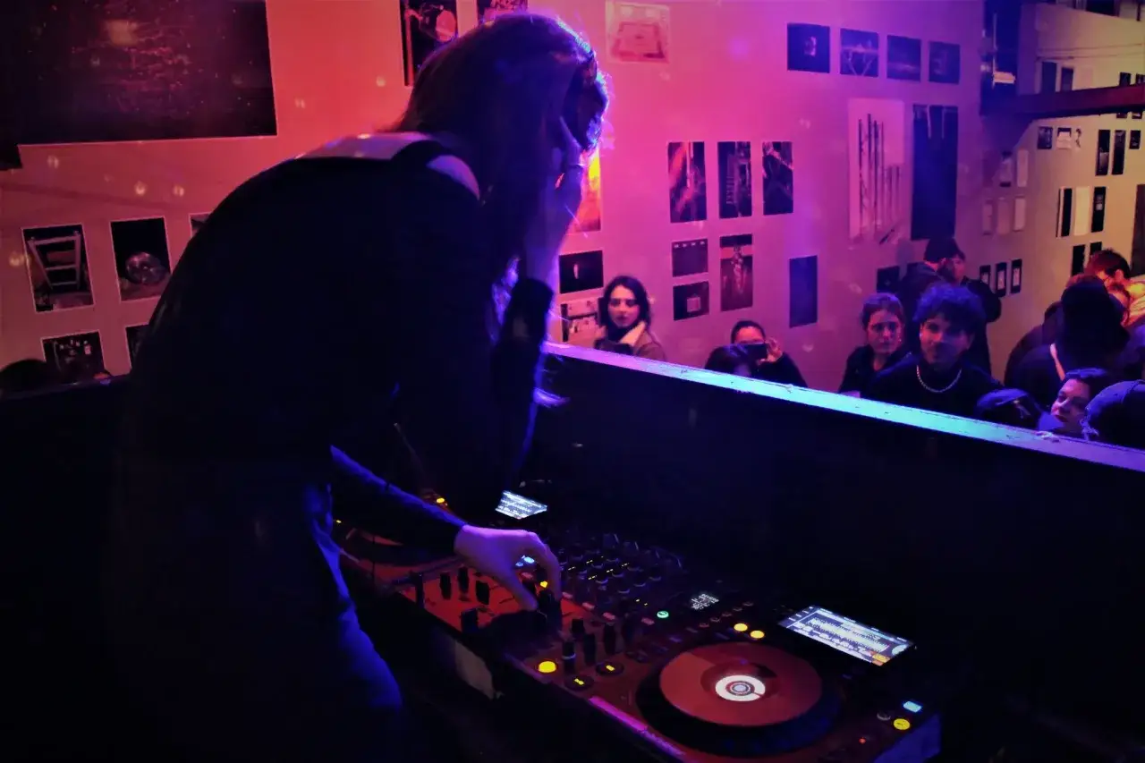 Une dj fille mixe dans une ambiance tamisée, la foule danse derrière elle.