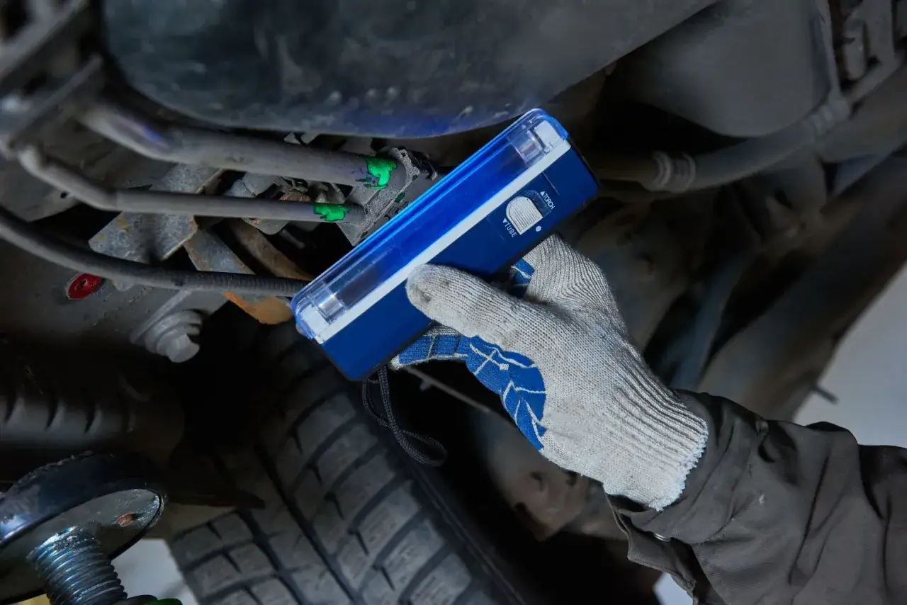 Gloved hand uses a UV light to find a freon leak on car AC lines. Green dye highlights the leak, showing how to fix freon leak in car.
