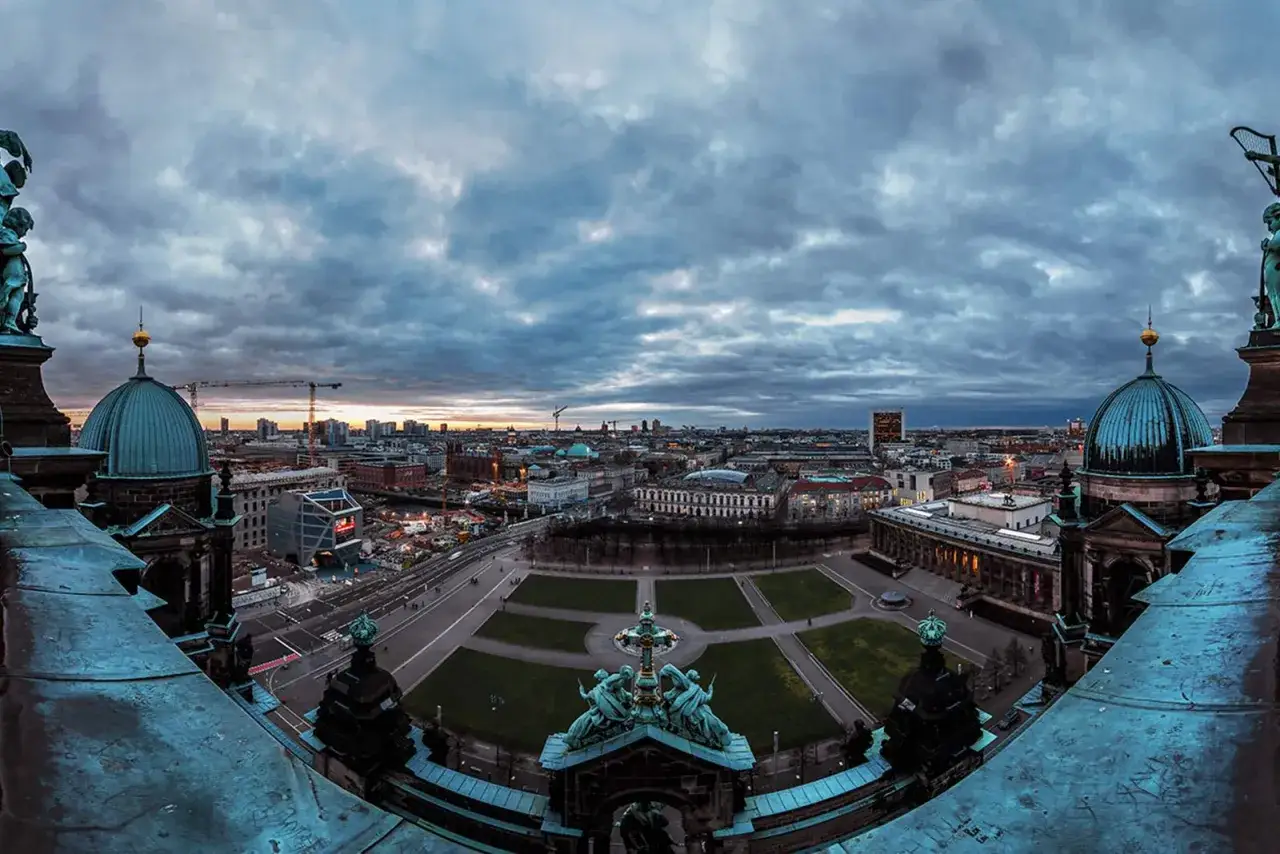 Blick vom Berliner Dom über die Stadt bei Dämmerung. Die Kuppeln des Doms dominieren den Vordergrund, während sich die Skyline Berlins unter einem dramatischen Wolkenhimmel erstreckt.