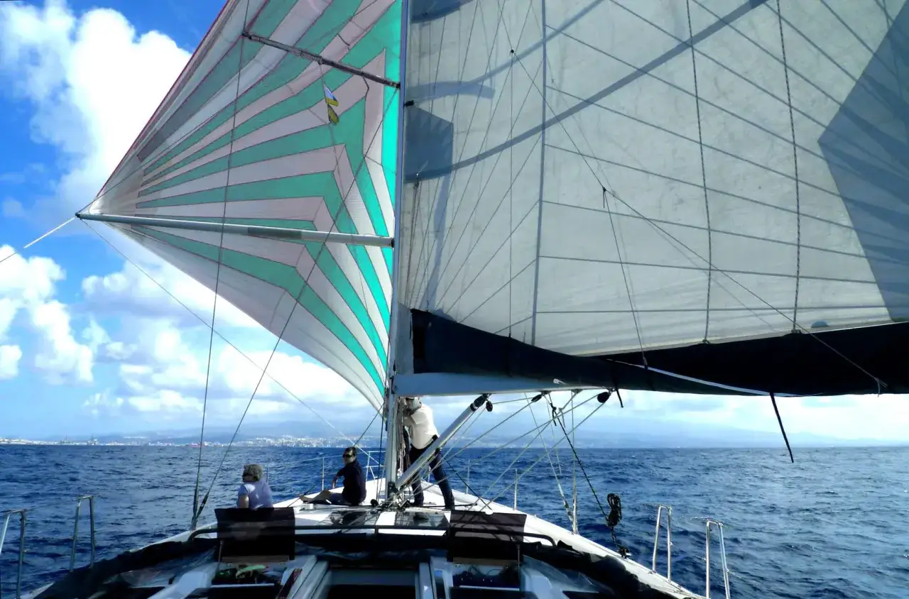 Vela colorata e bianca gonfie al vento, con persone a bordo. Si sta per mettersi alla cappa, godendosi il mare blu e il cielo nuvoloso.