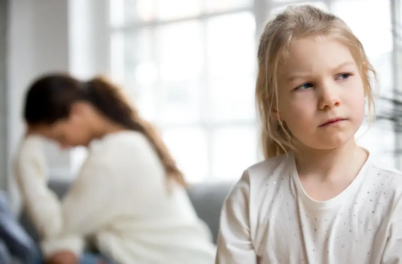 Smutna dziewczynka patrzy w bok, podczas gdy jej mama, zgarbiona, ukrywa twarz w dłoniach. To obrazek ilustrujący problem: parentyfikacja co to.
