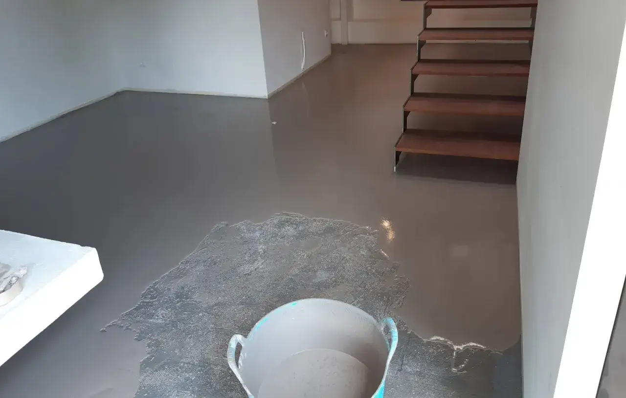 Un cubo con material grisáceo cerca de un **pavimento continuo hormigón** recién aplicado. Escaleras de madera al fondo.