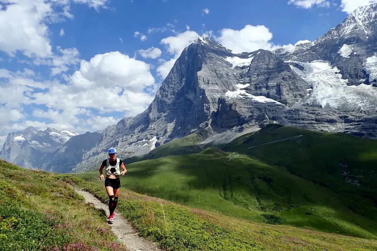 Une femme en plein effort de rando course sur un sentier de montagne, avec des sommets enneigés en toile de fond.