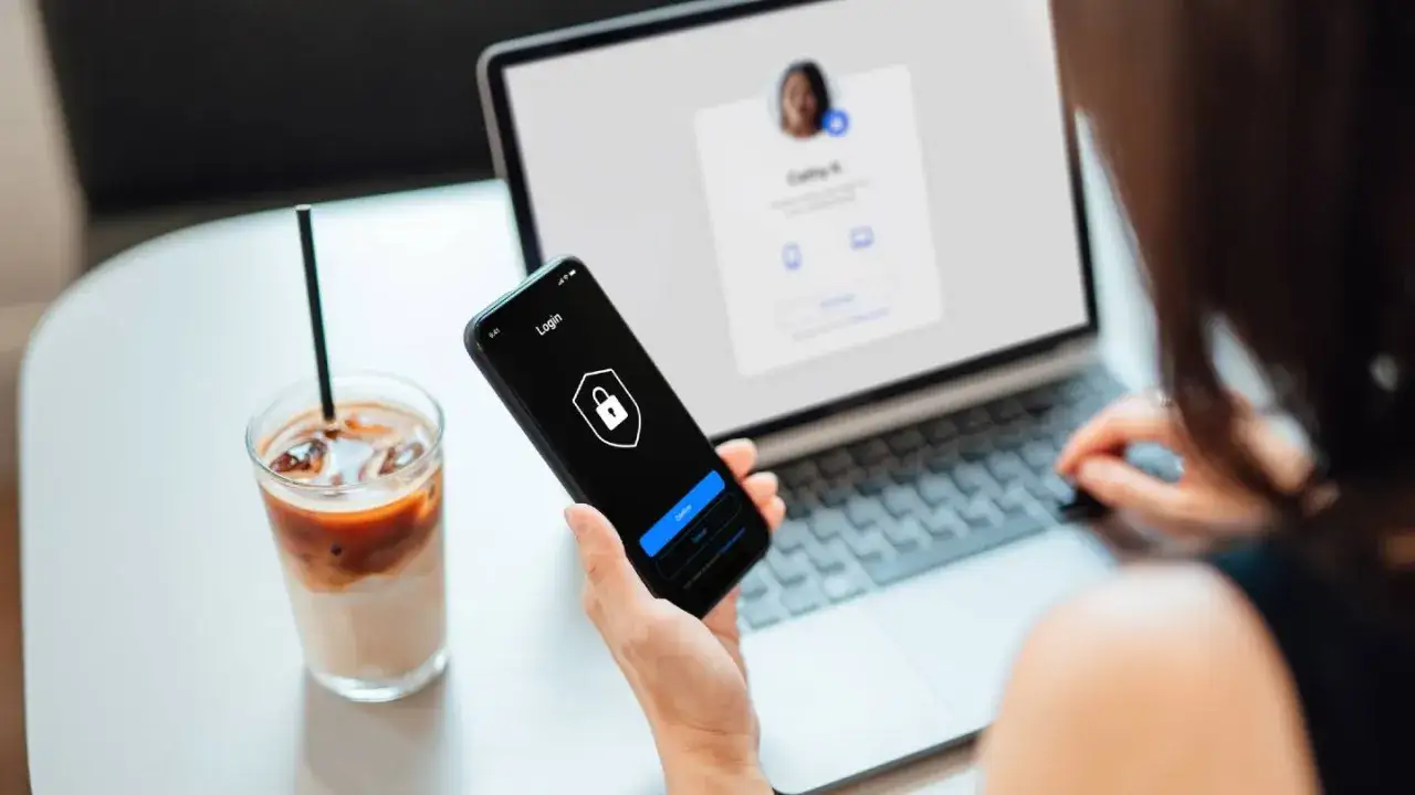 Kobieta używa telefonu z ikoną kłódki, sugerującą vpn na telefon. Na laptopie widoczny ekran logowania.
