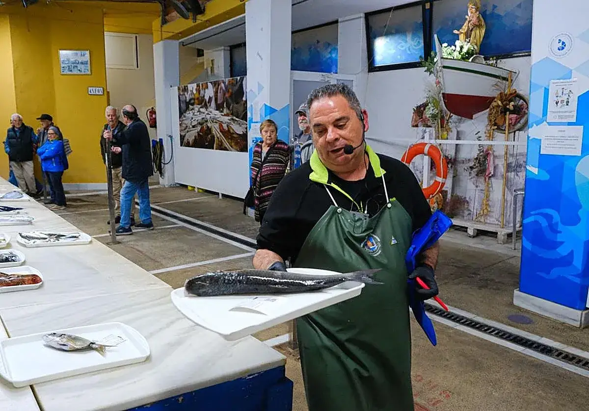 Hombre con delantal verde muestra un pescado. Detr&aacute;s, gente observa puestos con pescado, quiz&aacute;s en una subasta o mercado, evitando la pesca furtiva.