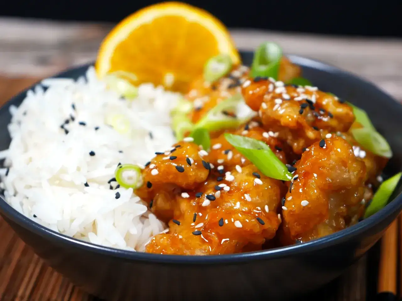 En skål med fluffigt ris och krispig, glaserad orange chicken, toppad med sesamfrön och salladslök. Ett perfekt recept på orange chicken!