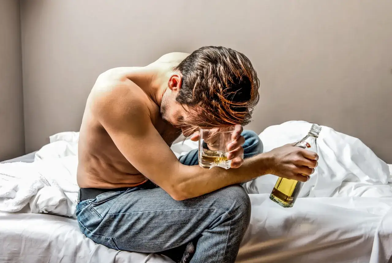 Mężczyzna siedzi na łóżku, trzymając szklankę z alkoholem i butelkę. Jego postawa sugeruje zmęczenie i potrzebę detoksykacji.