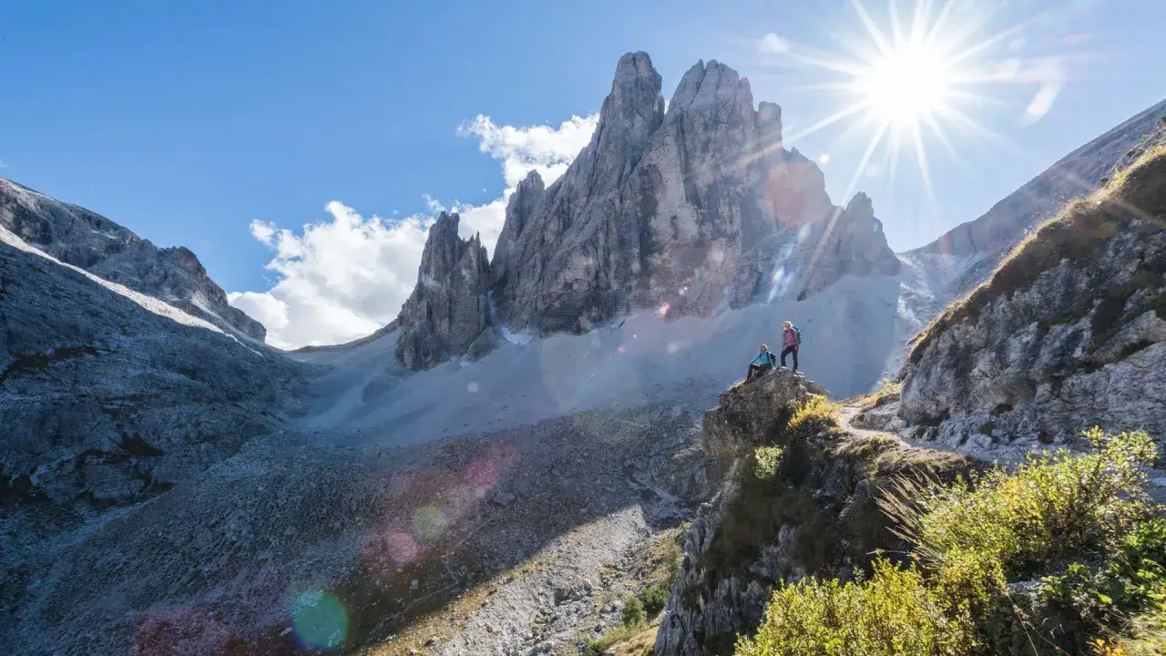 Zwei Wanderer genießen die atemberaubende Aussicht auf die majestätischen Dolomiten Berge. Die Sonne strahlt hell über den zerklüfteten Gipfeln.