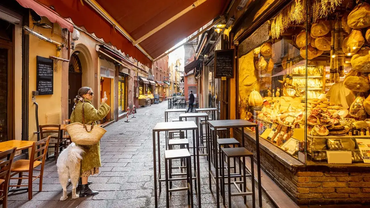 En kvinna med hund tar en bild p&aring; en charmig gata i Bologna, Italien, kantad av delikatesser och uteserveringar.