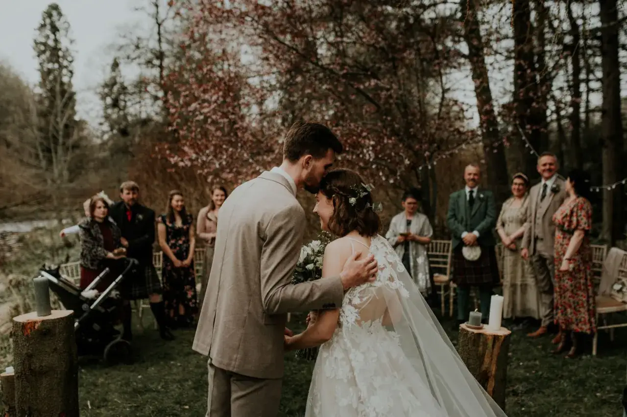 Ein Brautpaar k&uuml;sst sich bei einer romantischen **veganen Hochzeit** im Freien. G&auml;ste stehen im Hintergrund.