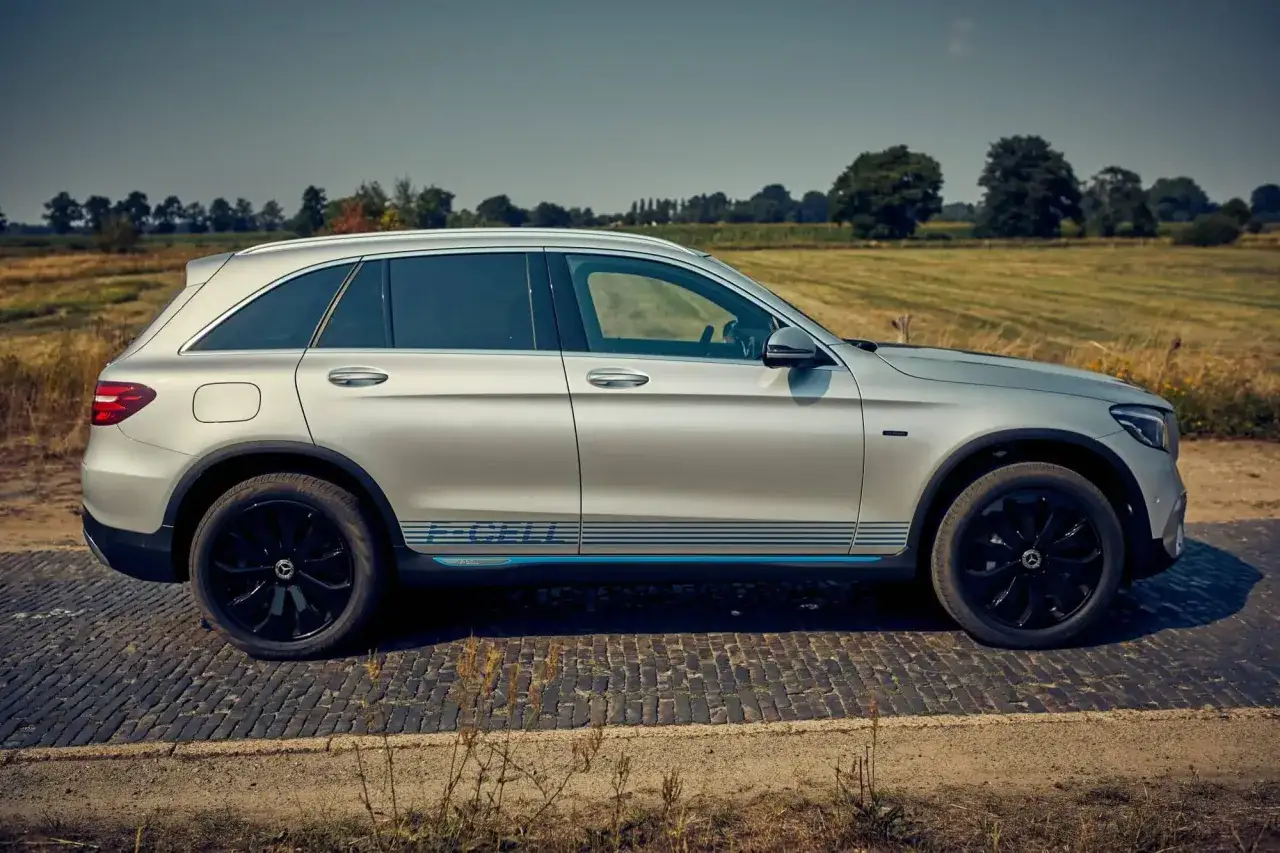 Voiture argentée, un **dimension GLC coupé** avec des bandes bleues sur le flanc, garée sur une route pavée devant un paysage champêtre.