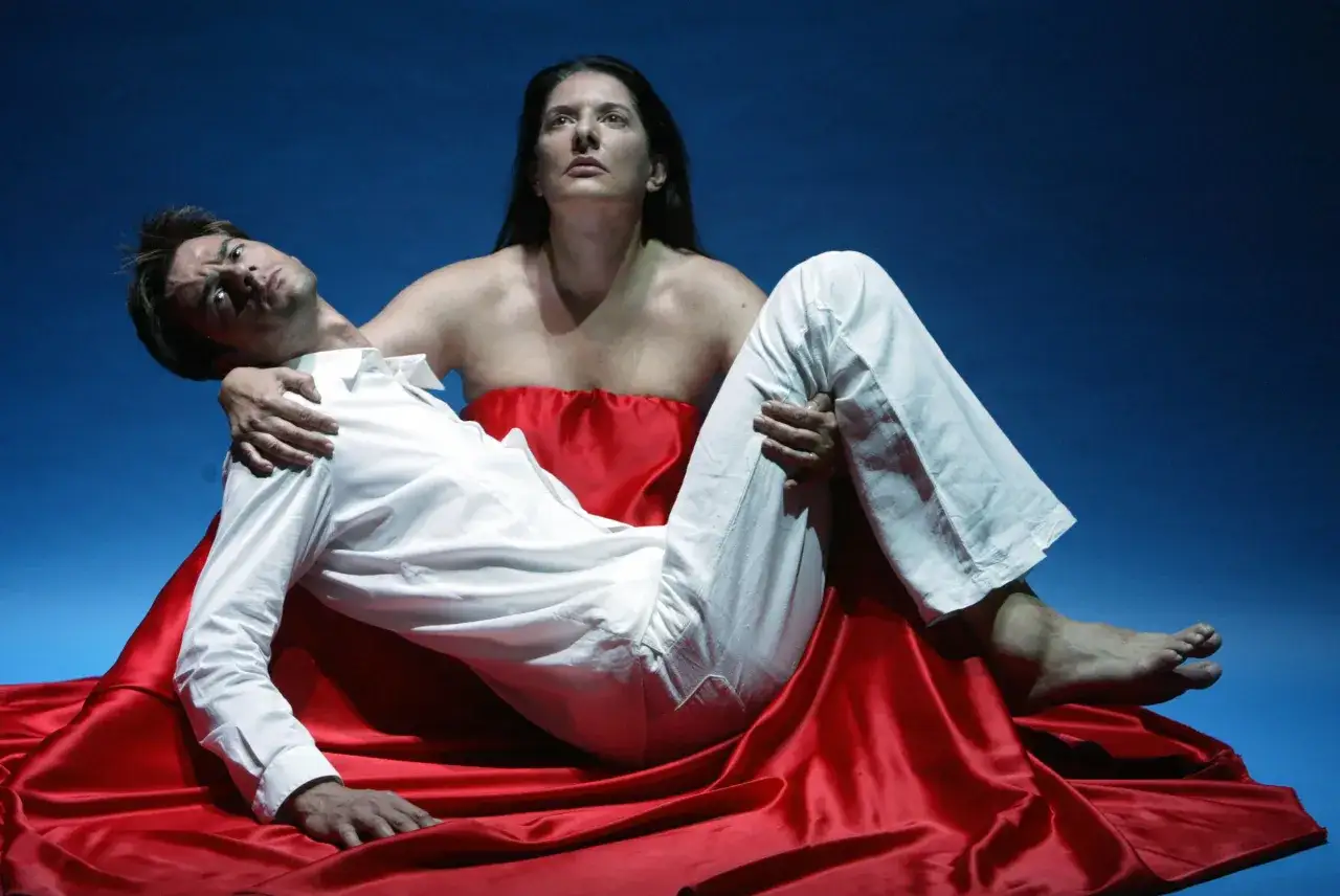 Kobieta w czerwonej draperii trzyma mężczyznę w białej koszuli. Scena jak z dramatycznego performance'u, co to za emocje?