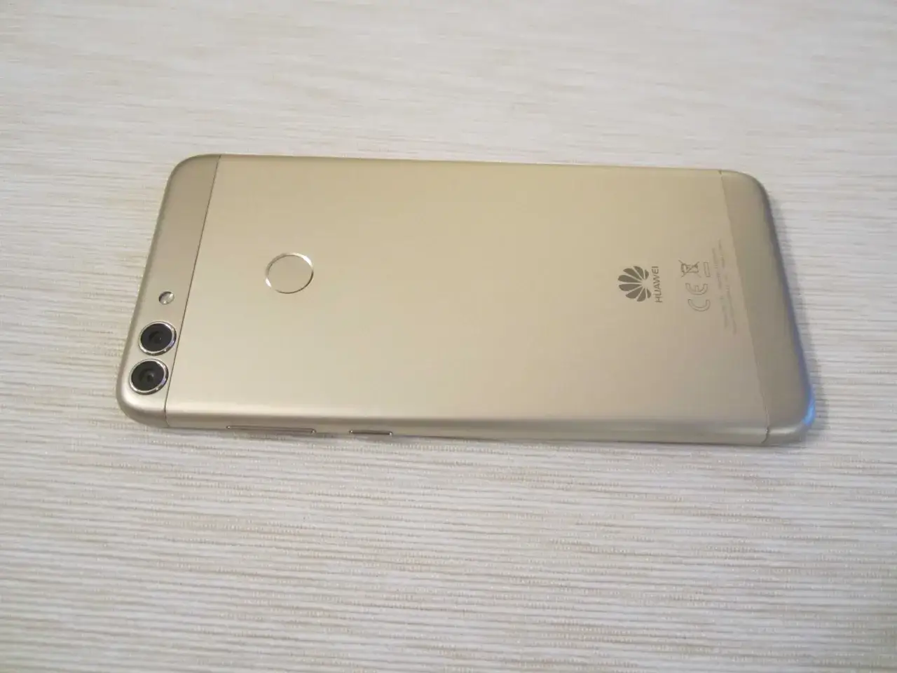 Złoty Huawei P Smart FIG-LX1 z podwójnym aparatem i czytnikiem linii papilarnych leży na jasnym, teksturowanym tle.