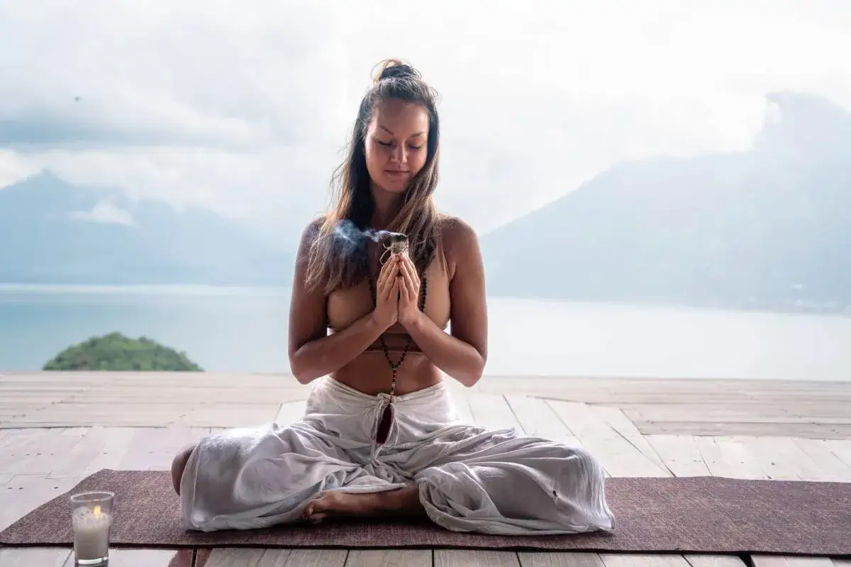 Mujer meditando en posición de loto, con las manos juntas y humo ascendiendo. Un sankalpa que es una intención profunda, se manifiesta en este momento de paz junto al lago.