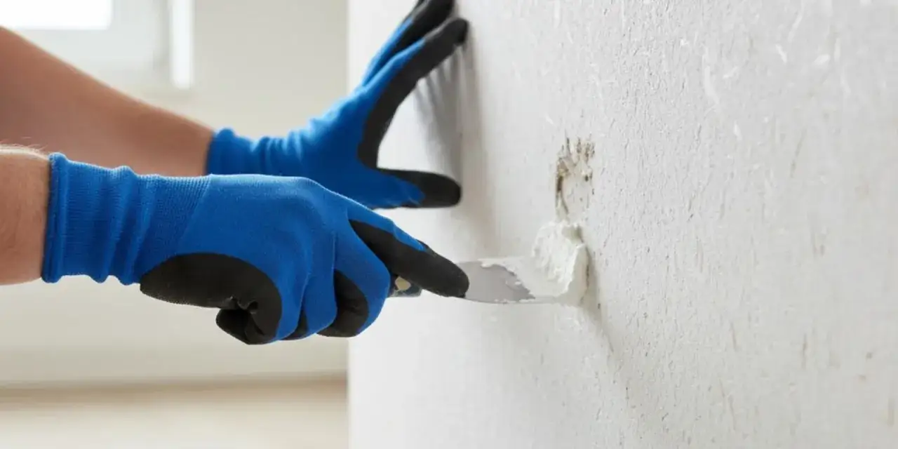 Hände mit blauen Handschuhen bessern eine beschädigte Stelle an einer Raufasertapete mit Spachtelmasse aus.