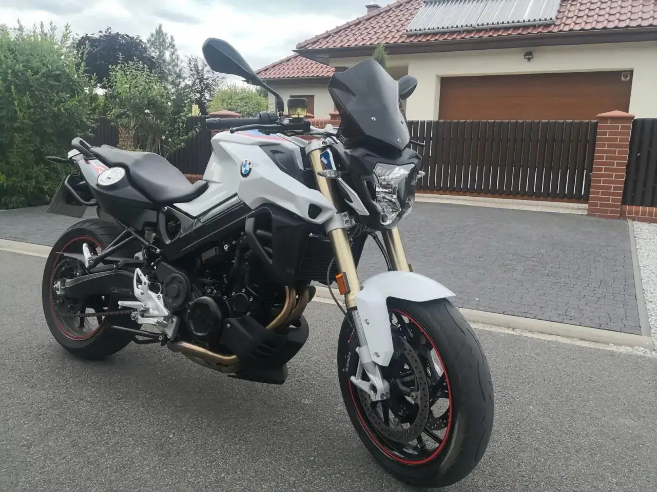 Biały motocykl BMW F 800 z czarną szybką i czerwonymi akcentami na felgach, zaparkowany przed domem.