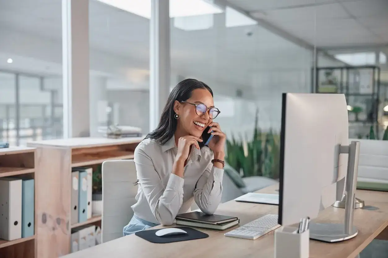 Donna sorridente al telefono in ufficio, mentre lavora al computer. Chiss&agrave; quanto guadagna una segretaria al mese con un lavoro cos&igrave; dinamico.