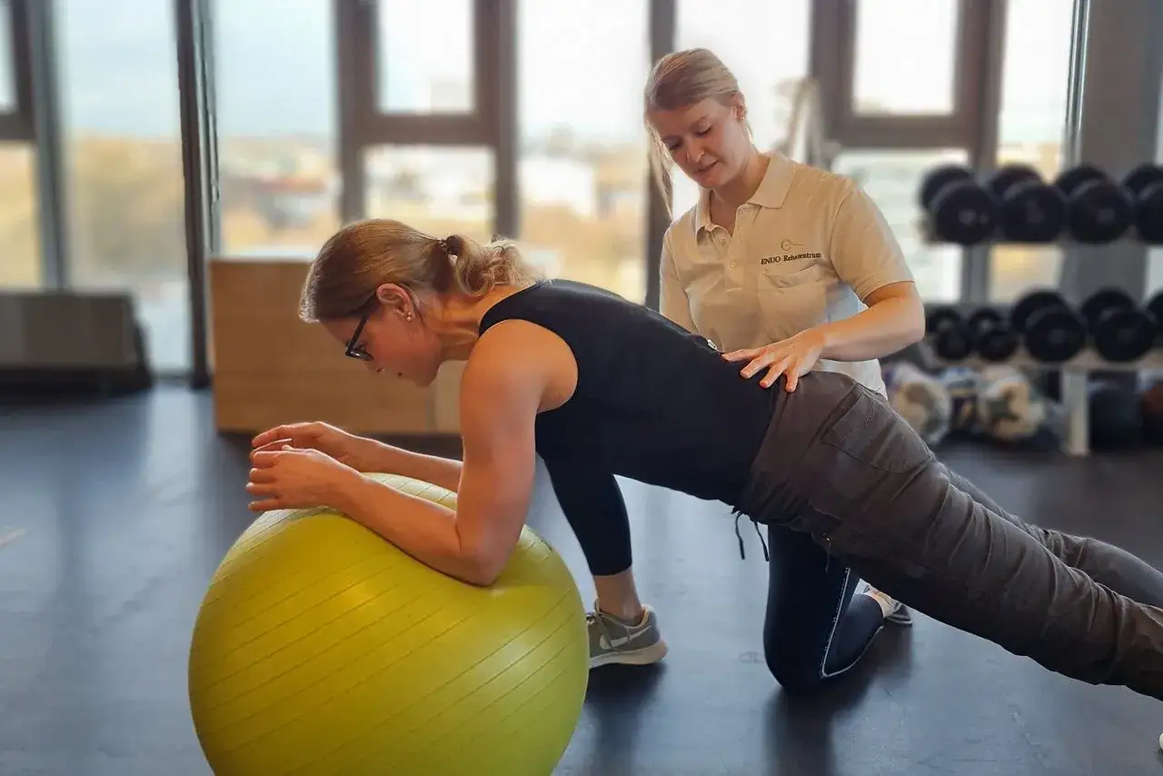 Die Physiotherapeuten unterstützen eine Frau bei einer Plank-Übung auf einem Gymnastikball.