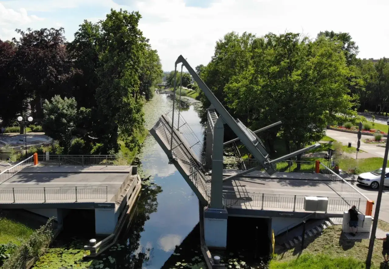 Podniesiony most zwodzony w Nowym Dworze Gdańskim, nad kanałem otoczonym zielenią.