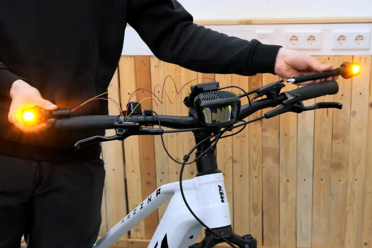 Person h&auml;lt einen leuchtenden Blinker am Fahrradlenker. Kabel verbinden ihn mit einem Scheinwerfer.