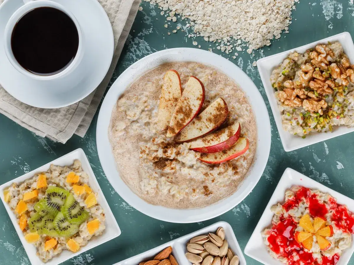 Un delicioso desayuno con porridge de avena, caf&eacute; y frutas. El porridge principal tiene rodajas de manzana y canela. Otros cuencos muestran avena con kiwi, naranja, nueces, pistachos y mermelada.