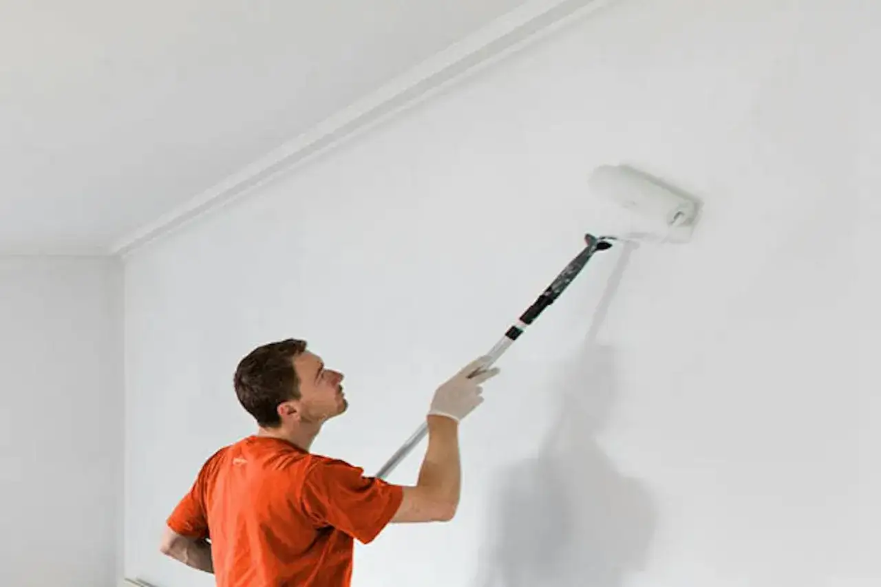 Man i orange tr&ouml;ja anv&auml;nder en l&aring;ng roller f&ouml;r att m&aring;la grund p&aring; en vit v&auml;gg.