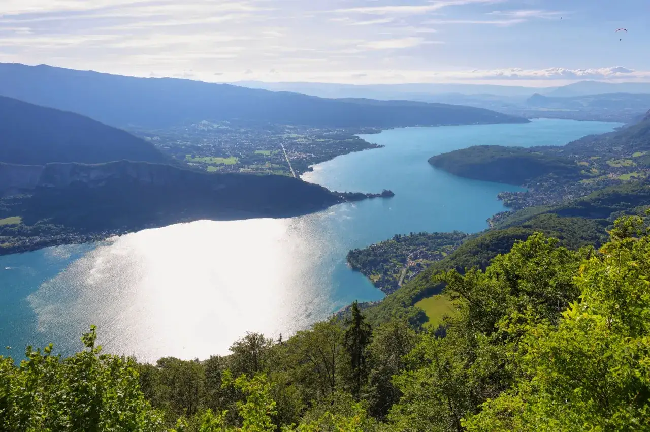 Vue panoramique sur le lac d'Annecy, entouré de montagnes verdoyantes. Deux parapentistes volent dans le ciel bleu.