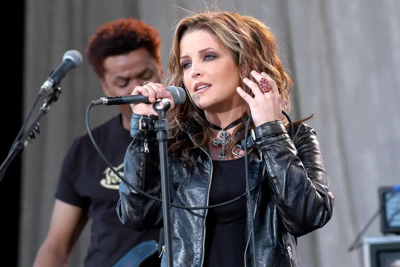 Lisa Marie Presley śpiewa na scenie, ubrana w skórzaną kurtkę i naszyjnik z krzyżem.