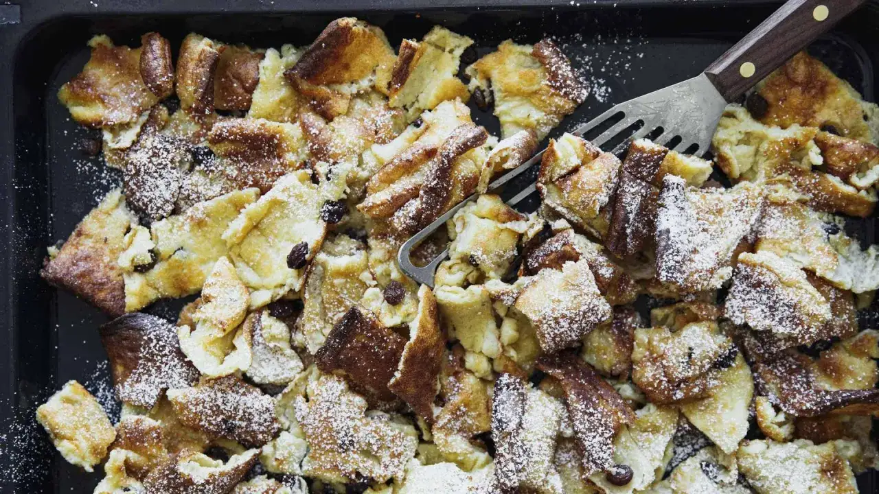 Kaiserschmarrn aus dem Backofen auf einem Blech, mit Puderzucker bestäubt und Rosinen. Ein Gabel-Schaber liegt bereit.